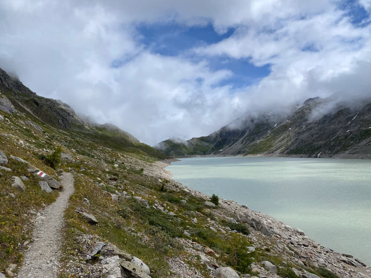 Wanderweg über einem milchig-grünen Stausee, Wolken an den Flanken